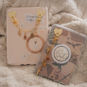 Op de afbeelding: Twee telefoonhoesjes met decoratieve gouden kettingen en bedels. Eén hoesje is lichtroze met madeliefjesstickers en de tekst "Winglen & High Ls." De andere is transparant met een madeliefjes-popsocket en een vintage print.