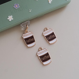 Puede incluir: Encantos dorados de taza de café con la palabra "coffee" en fuente blanca. Los encantos son marrones y blancos, con un borde dorado y un pequeño lazo en la parte superior. Un encanto está unido a un agarre transparente para el teléfono.
