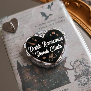 Puede incluir: Un soporte para teléfono con forma de corazón negro con el texto "Dark Romance Book Club" en escritura blanca. El soporte tiene un borde festoneado blanco y detalles dorados, incluyendo esposas y estrellas. También se ve un soporte para teléfono en forma de corazón plateado.