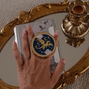 Peut inclure: Un support de téléphone circulaire avec un fond bleu foncé orné d'un dragon doré, d'un croissant de lune, d'étoiles et de nuages violet clair. Le support présente un bord doré festonné et est fixé à une coque de téléphone transparente.