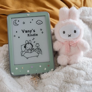 Op de afbeelding: Een mintgroene Kindle e-reader met een bloemenontwerp en de tekst "Vany's Kindle" met een cartoon illustratie. Een roze pluche konijn speelgoed met een strik ligt naast de e-reader op een witte, zachte deken.