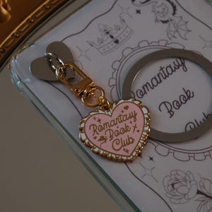 Puede incluir: Un llavero en forma de corazón rosa y dorado con el texto "Romantasy Book Club". El llavero tiene un cierre dorado y un accesorio en forma de corazón plateado. El fondo presenta un diseño decorativo con una corona y un dragón.