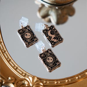 May include: Three gold-coloured tarot card earrings with black enamel detailing. The earrings feature celestial designs of the sun, moon, and stars, with the words "THE MAGICIAN" and "THE MOON" on the cards. The earrings are displayed on a reflective surface.