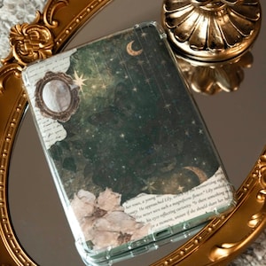 Puede incluir: Una funda transparente para teléfono con un diseño floral y verde oscuro, con lunas y estrellas. La funda descansa sobre un espejo con marco dorado, con un candelabro dorado en el fondo.
