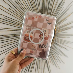 K&ouml;nnte beinhalten: Transparente Handyh&uuml;lle mit einem rosa-wei&szlig;en Karomuster, verziert mit Halloween-Stickern. Die Sticker zeigen ein K&uuml;rbisgew&uuml;rzglas, einen Geist und das Wort "Boo!".