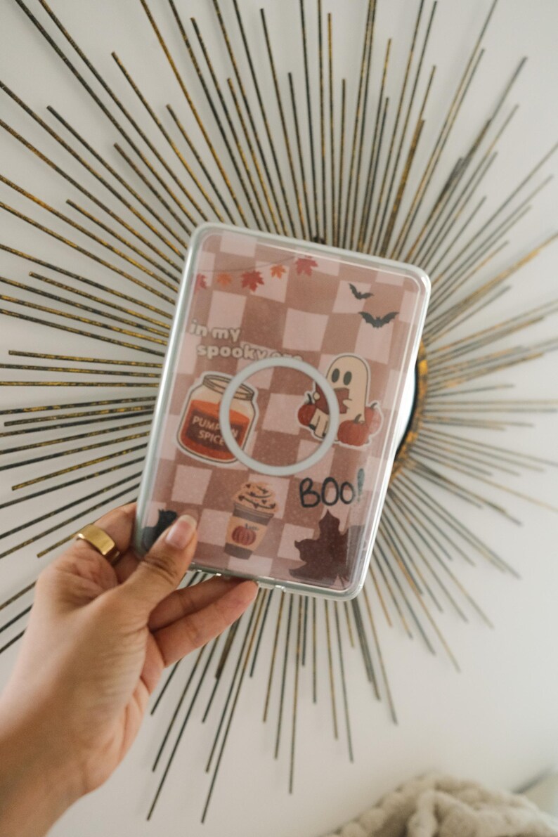 K&ouml;nnte beinhalten: Transparente Handyh&uuml;lle mit einem rosa-wei&szlig;en Karomuster, verziert mit Halloween-Stickern. Die Sticker zeigen ein K&uuml;rbisgew&uuml;rzglas, einen Geist und das Wort "Boo!".