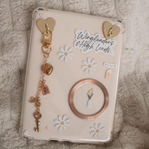 Könnte beinhalten: Eine beige Tablet-Hülle mit goldfarbenen herzförmigen Akzenten und einem Anhänger. Der Anhänger enthält ein Herz, eine Rose und einen Schlüssel. Die Hülle ist mit Gänseblümchen-Aufklebern und dem Text "Wingleaders & High Lords" verziert.
