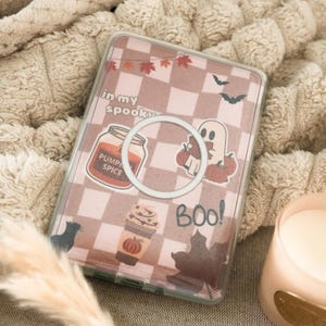 Op de afbeelding: Transparante telefoonhoes met een bruin en wit geblokt patroon en Halloween-stickers. De stickers bevatten een spook, pompoenen, een pot pompoenkruiden en de woorden "in my spooky" en "BOO!"