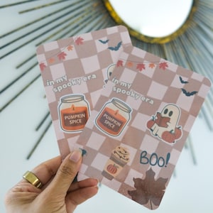 May include: Two rectangular cards with a light brown checkered pattern and autumn illustrations. The cards feature text that reads "in my spooky era" and "Pumpkin Spice". One card has a ghost reading a book and the word "BOO!"
