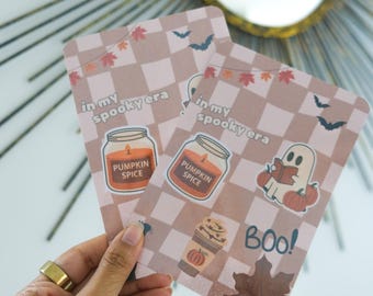 May include: Two rectangular cards with a light brown checkered pattern and autumn illustrations. The cards feature text that reads "in my spooky era" and "Pumpkin Spice". One card has a ghost reading a book and the word "BOO!"