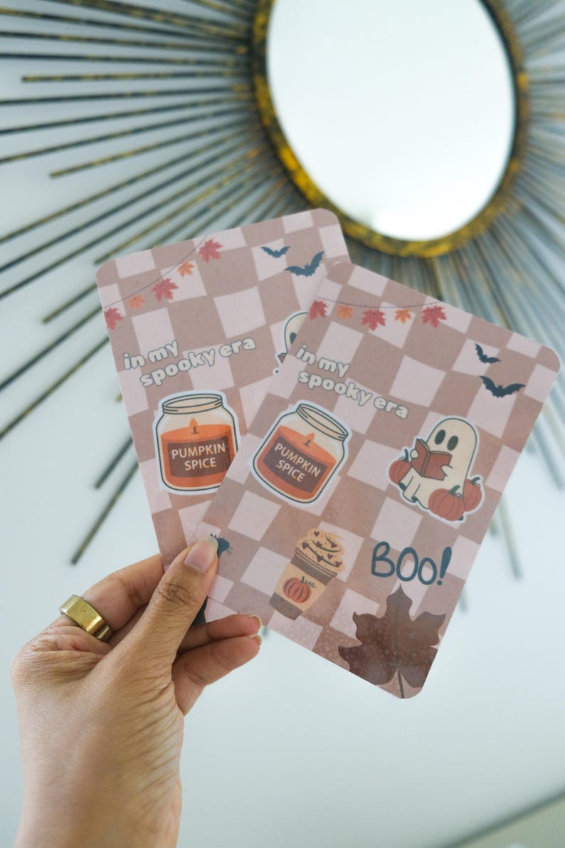 K&ouml;nnte beinhalten: Zwei rechteckige Karten mit hellbraunem Karomuster und herbstlichen Illustrationen. Die Karten zeigen den Text "in my spooky era" und "Pumpkin Spice". Eine Karte hat einen Geist, der ein Buch liest, und das Wort "BOO!"
