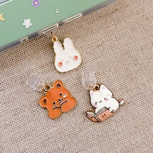 Op de afbeelding: Drie emaille bedels met goudkleurige bevestigingen. De bedels tonen een witte konijn met roze wangen, een bruine beer en een witte kat die een boek leest. De bedels zijn bevestigd aan doorzichtige oorbel sluitingen.
