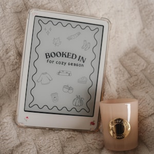 May include: An e-reader with a clear case displays the text "BOOKED IN for cozy season" with fall-themed illustrations. A light pink candle with a gold emblem sits beside the e-reader, creating a cozy aesthetic.
