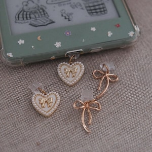 May include: A selection of earrings and charms. Two heart-shaped charms with gold edging, white enamel, and bow designs are shown. A pair of asymmetrical bow earrings are also displayed. A green e-reader case with floral details is in the background.