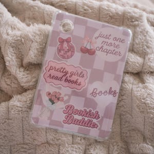 May include: A clear phone case with a pink and white chequered pattern. The case has stickers with text and illustrations, including a disco ball, cherries, and the phrases "pretty girls read books" and "just one more chapter."