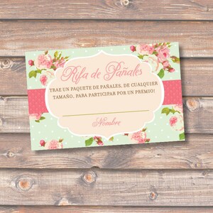 May include: A floral baby shower raffle card with the text "Rifa de Pañales". The card features a light green background with white polka dots, a pink stripe, and a decorative frame with pink roses. The card is on a wooden surface.