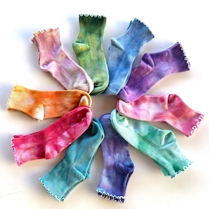 May include: A circular arrangement of ten ribbed ankle socks, each featuring a unique tie-dye pattern in vibrant colors including pink, orange, yellow, green, blue, and purple. Each sock has a distinct ruffled lettuce-edge trim at the cuff.
