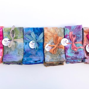 May include: Six folded cloth napkins featuring tie-dye patterns in vibrant shades of pink, green, blue, orange, and purple. Each napkin is tied with a matching fabric ribbon and includes a small circular white tag with the text Allison Figs.