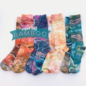 May include: A collection of colorful socks with a marbled pattern in shades of orange, brown, blue, and green. The socks are arranged on a white surface. The text "super soft BAMBOO" is displayed on a teal banner.