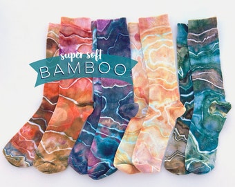 May include: A collection of colorful socks with a marbled pattern in shades of orange, brown, blue, and green. The socks are arranged on a white surface. The text "super soft BAMBOO" is displayed on a teal banner.