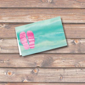 May include: A folded greeting card featuring a watercolor design of pink flip-flops on a turquoise background. The card is set against a rustic wooden surface, evoking a beach or summer theme.