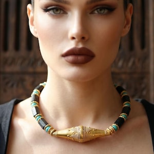 May include: A close-up of a woman wearing a gold and black necklace with turquoise accents. The necklace has a central gold pendant with intricate detailing. The woman has dark hair and makeup, and is wearing a black top.