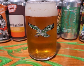 May include: A clear glass beer tumbler filled with amber-colored beer and a foamy head. The glass features a green and black graphic of an eagle holding a football. Canned beers are visible in the background.