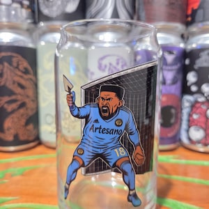 May include: Clear glass beer can shaped glass featuring a soccer player graphic. The player is wearing a blue uniform with the word "Artesano" and the number "18". The player is holding a trowel in front of a soccer net.
