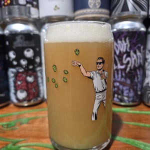 May include: A glass of beer with a white foamy head. The glass features a cartoon illustration of a man in sunglasses throwing hops. The beer is a light amber color. Cans of beer are visible in the background.