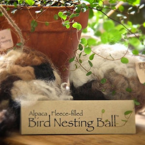 May include: Two brown and white alpaca fleece-filled bird nesting balls. The balls are handmade and have a natural, rustic look. The text on the tag reads "Alpaca, Fleece-filled Bird Nesting Ball".