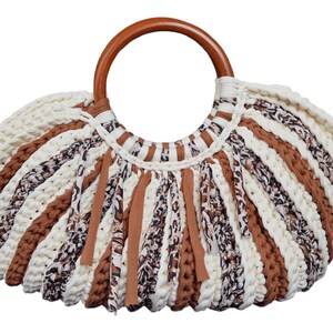 Könnte beinhalten: Eine handgefertigte Häkeltasche mit Holzgriff. Die Tasche hat abwechselnde Streifen aus Creme, Braun und gemustertem Stoff. Braune Stoffstreifen hängen vom unteren Rand. Der Griff ist ein großer, glatter Holzring.