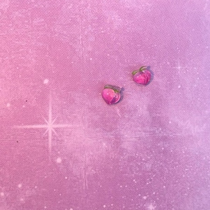 Pode incluir: Dois brincos em forma de pêssego com hastes e folhas verdes. Os brincos são rosa e têm um acabamento brilhante. Eles estão em um fundo rosa.