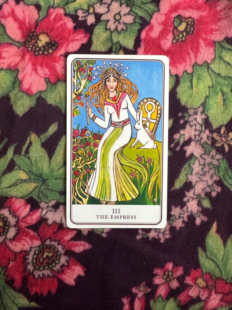以下が含まれることがあります： 女帝のイラストが描かれたタロットカード。カードには、長い金髪で、白と緑のドレスを着て、白いウサギと一緒に玉座に座っている女性が描かれています。カードには「III THE EMPRESS」と書かれています。