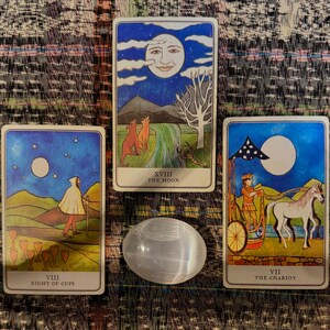 以下が含まれることがあります： カラフルなイラストが描かれた3枚のタロットカードと、磨かれた楕円形のセレナイトクリスタル。カードには月、人物、動物が登場するシーンが描かれています。カードには「Eight of Cups」、「The Moon」、「The Chariot」と書かれています。