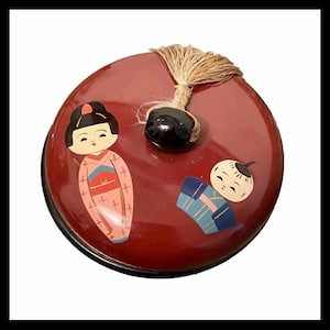 May include: A round, red lacquered box with a black base and a tassel. The lid features two Japanese-style illustrations: a woman in a pink kimono and a boy in a blue outfit. The box has a black bead on top.