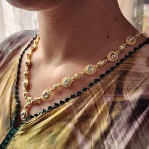 Peut inclure: Un collier doré orné de pierres précieuses rondes et transparentes, monté sur une chaîne délicate. Le collier est porté sur un haut à motifs avec une bordure verte. Les pierres sont serties dans un motif circulaire.