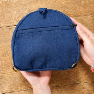 May include: A semi-circular dark blue fabric pouch featuring a horizontal zipped pocket across the front, a small top tab, and two brass-toned metal rings attached to the bottom corners for strap attachment, showcasing a textured weave.