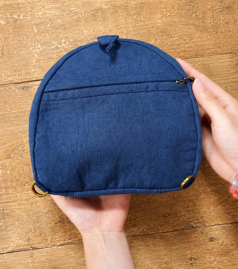 May include: A semi-circular dark blue fabric pouch featuring a horizontal zipped pocket across the front, a small top tab, and two brass-toned metal rings attached to the bottom corners for strap attachment, showcasing a textured weave.