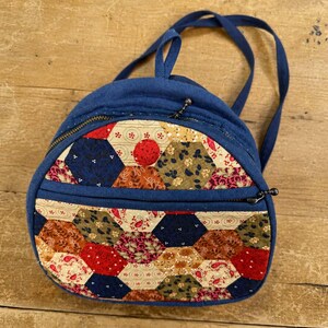 May include: A small backpack featuring a solid navy blue fabric body and a front panel decorated with a patchwork of multicoloured hexagonal fabric pieces in shades of red, blue, green, and cream. It includes two zippered compartments and straps.