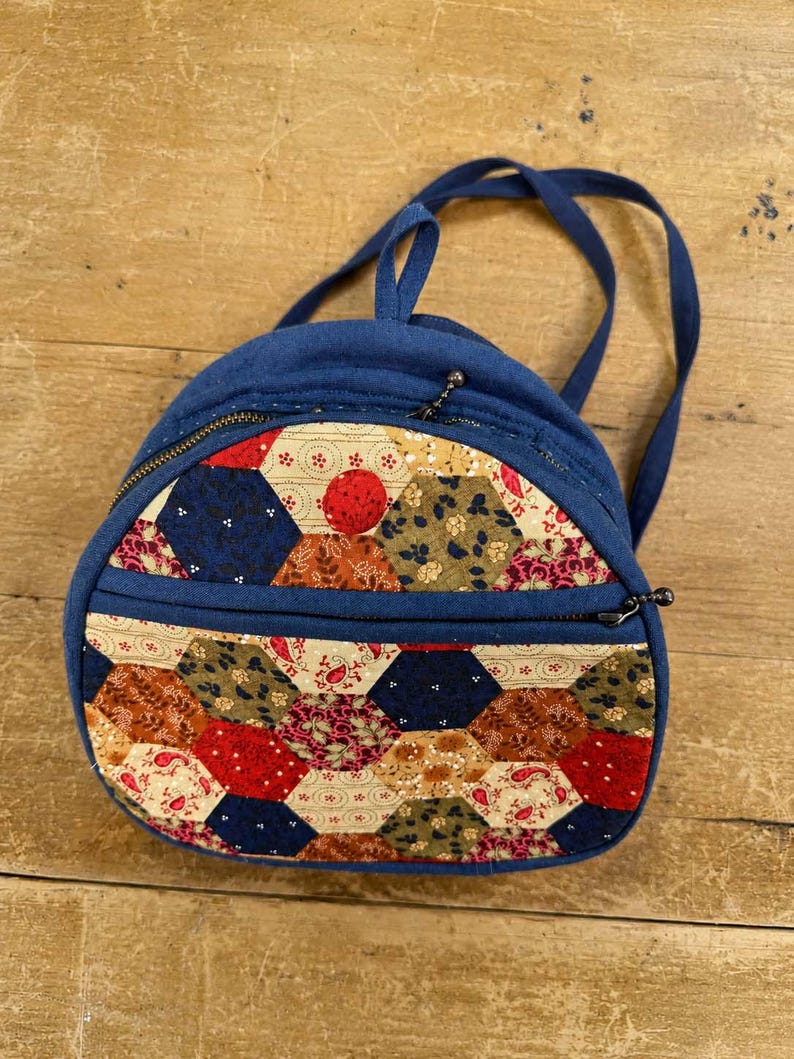 May include: A small backpack featuring a solid navy blue fabric body and a front panel decorated with a patchwork of multicoloured hexagonal fabric pieces in shades of red, blue, green, and cream. It includes two zippered compartments and straps.
