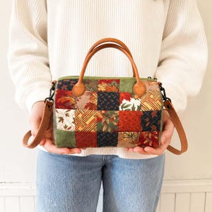 Puede incluir: Un bolso patchwork con asa y bandolera de cuero marrón. El bolso presenta cuadrados de tela en colores otoñales, incluyendo rojo, verde y dorado. El bolso es sostenido por una persona.