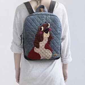 Puede incluir: Una mochila acolchada de color gris con un diseño bordado de un perro marrón y blanco en el panel frontal. La bolsa incluye correas de hombro de color gris oscuro, un asa superior y una pequeña etiqueta rectangular de marca en la esquina inferior.