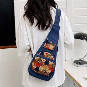 May include: A blue canvas crossbody sling bag featuring a patchwork hexagonal pattern in various floral and geometric prints on the front pocket and a smaller matching flap pocket with a gold-tone button closure and a vertical zipper.