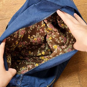 May include: A navy blue canvas bag with a zip closure, featuring an interior lining with a brown background and a floral pattern of pink roses and yellow flowers. The bag includes a circular metal zip pull and matching blue fabric straps.