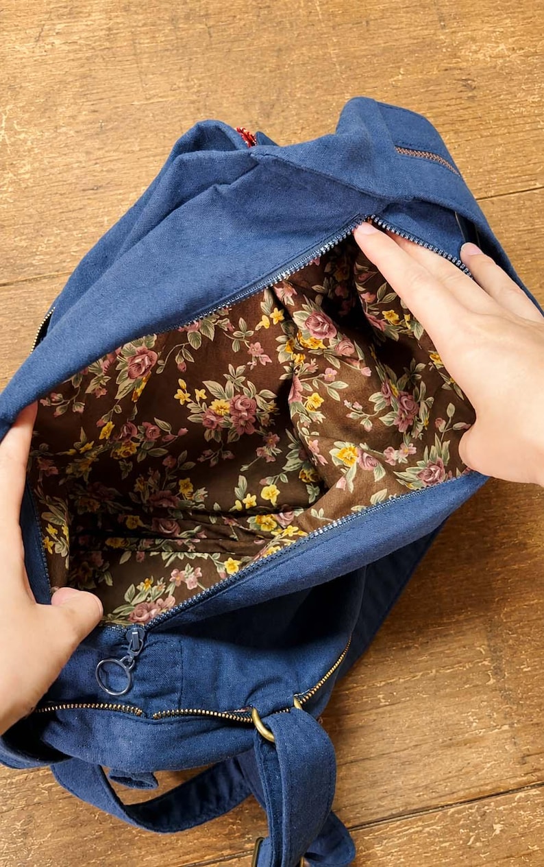 May include: A navy blue canvas bag with a zip closure, featuring an interior lining with a brown background and a floral pattern of pink roses and yellow flowers. The bag includes a circular metal zip pull and matching blue fabric straps.