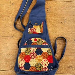May include: A blue canvas sling backpack featuring a teardrop shape, a single shoulder strap with a zip, and two exterior pockets. The pockets are decorated with a colourful patchwork pattern of fabric hexagons in shades of red, yellow, and green.