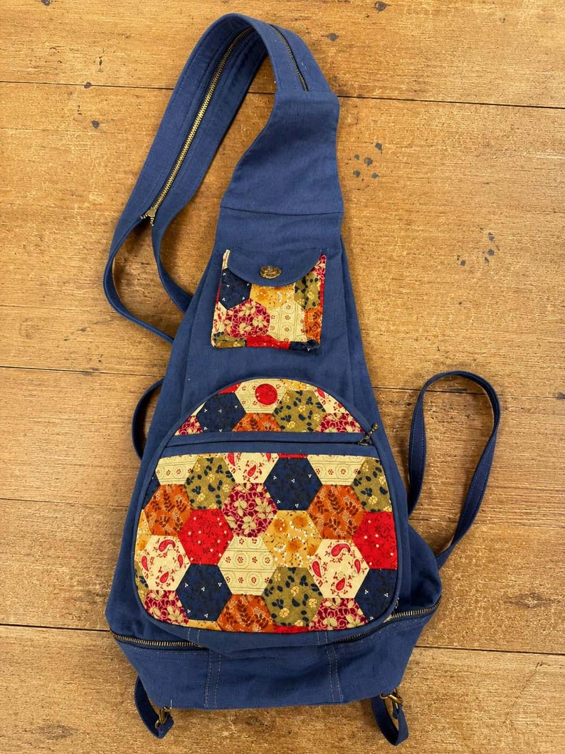 May include: A blue canvas sling backpack featuring a teardrop shape, a single shoulder strap with a zip, and two exterior pockets. The pockets are decorated with a colourful patchwork pattern of fabric hexagons in shades of red, yellow, and green.