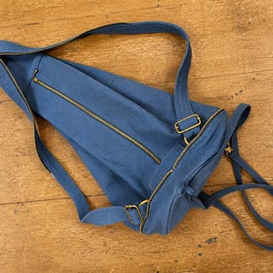 May include: A blue canvas backpack featuring a long diagonal zip closure across the main compartment, a smaller zipped pocket, and adjustable shoulder straps with gold-toned metal hardware. The bag has a cylindrical shape and a soft structure.