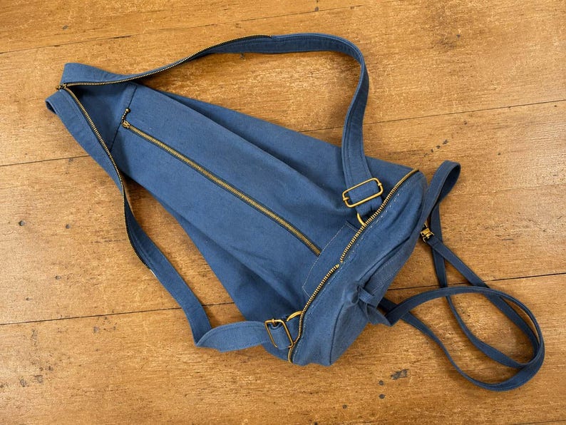 May include: A blue canvas backpack featuring a long diagonal zip closure across the main compartment, a smaller zipped pocket, and adjustable shoulder straps with gold-toned metal hardware. The bag has a cylindrical shape and a soft structure.
