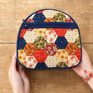 May include: A semi-circular zipped pouch featuring a patchwork design of multi-coloured fabric hexagons. The exterior displays various floral and paisley patterns in shades of red, navy blue, orange, and cream, finished with a solid navy blue trim.